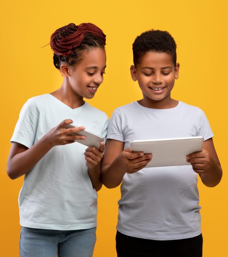 Gadgets addiction among kids concept. Cheerful black girl teenager with modern smartphone and boy with brand new digital tablet playing mobile games or using nice applications on yellow