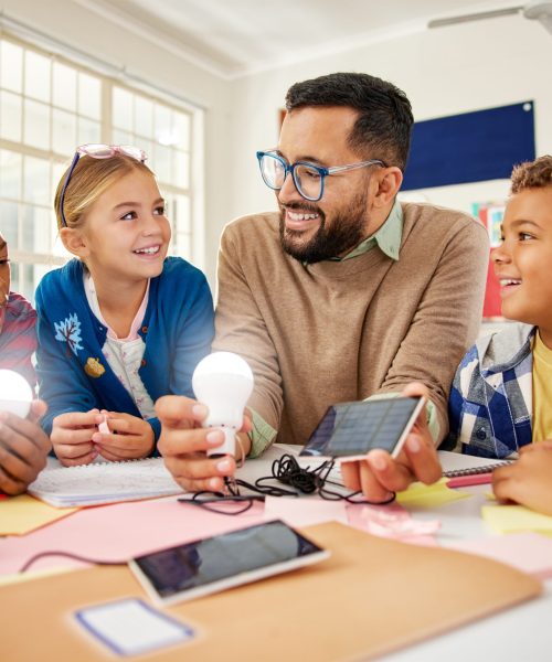 Happy male teacher with solar panel explaining pupil about solar energy with illuminated bulbs. Teacher in science classroom showing a solar panel to generate electricity. Smiling multiethnic primary children learning and experimenting about renewable energy sitting together around desks.