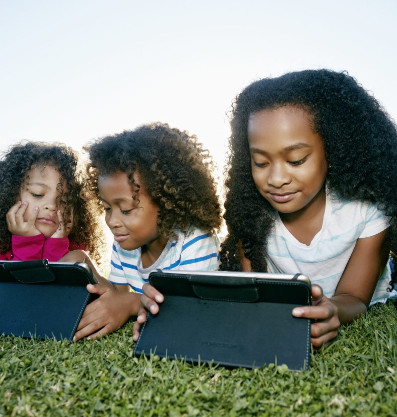 Young mixed race girl and her brother and sister lying on grass with two digital tablets, screen time